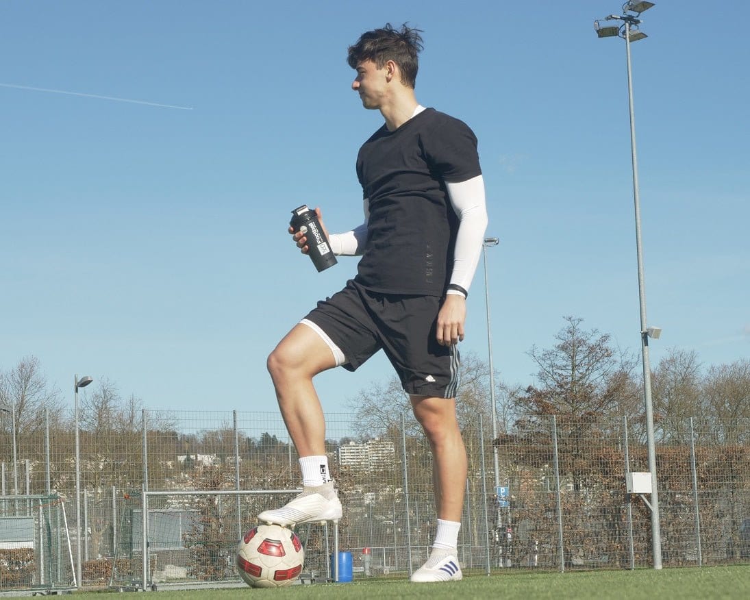 Ein Fussballer steht mit einem Shaker und Creatine360 auf dem Fussballplatz